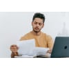 Man reading printed documents with his laptop in front of him
