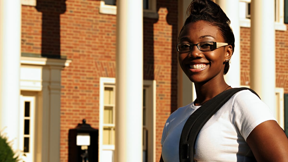 Black student with glasses smiling in front of a college building