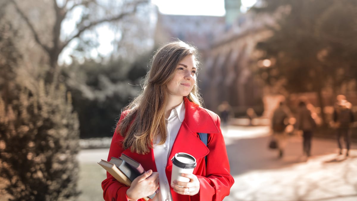 Student in a red coat holding books and a coffee