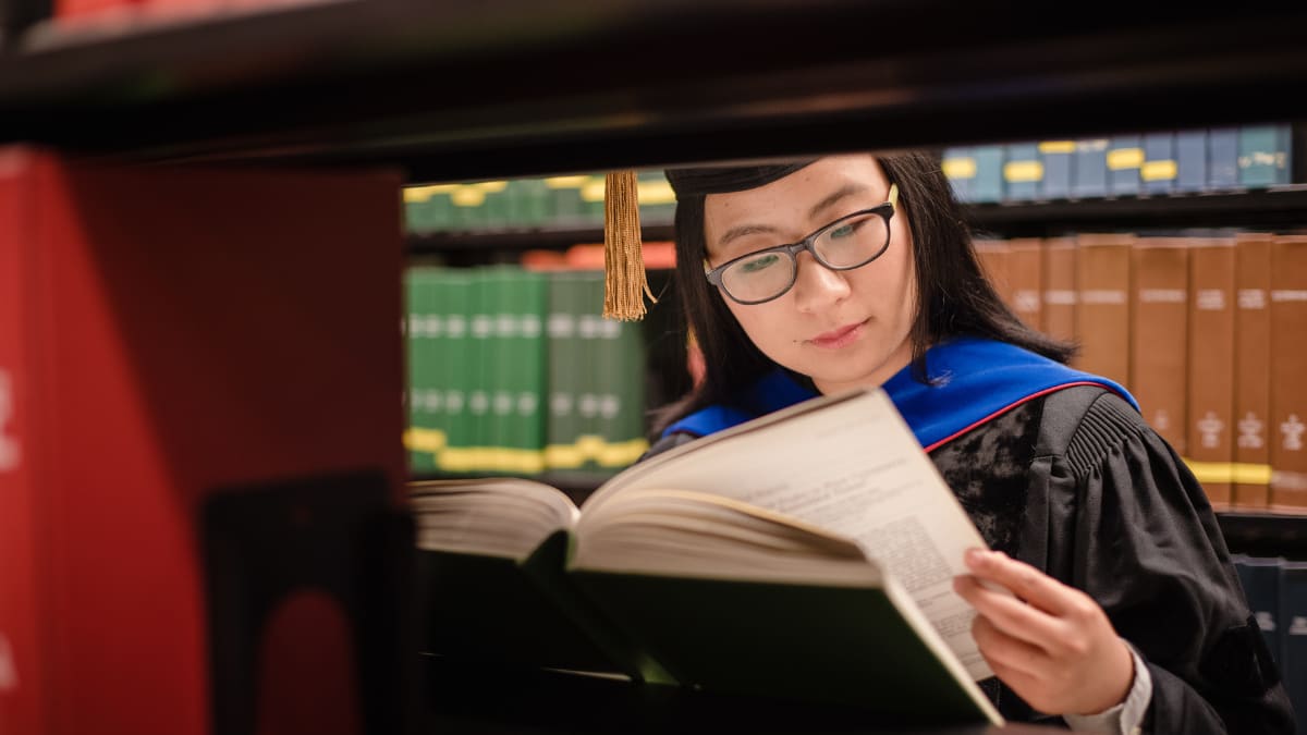 Graduate in cap and gown reading a book in the library
