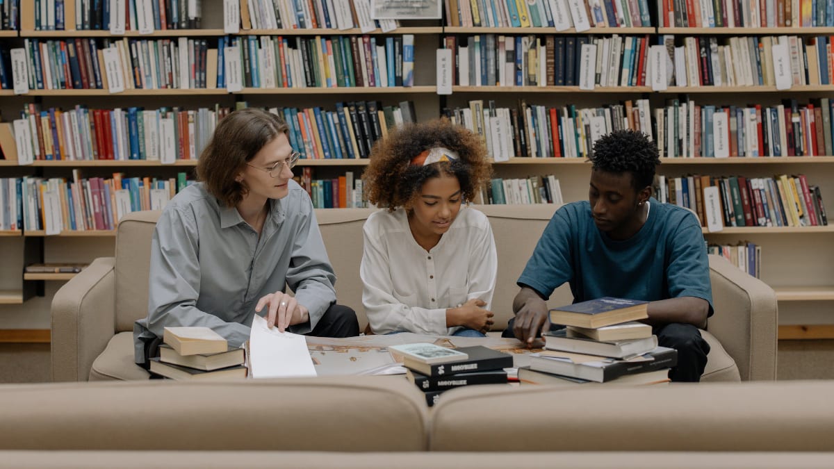 three students sitting in a library looking at a book together