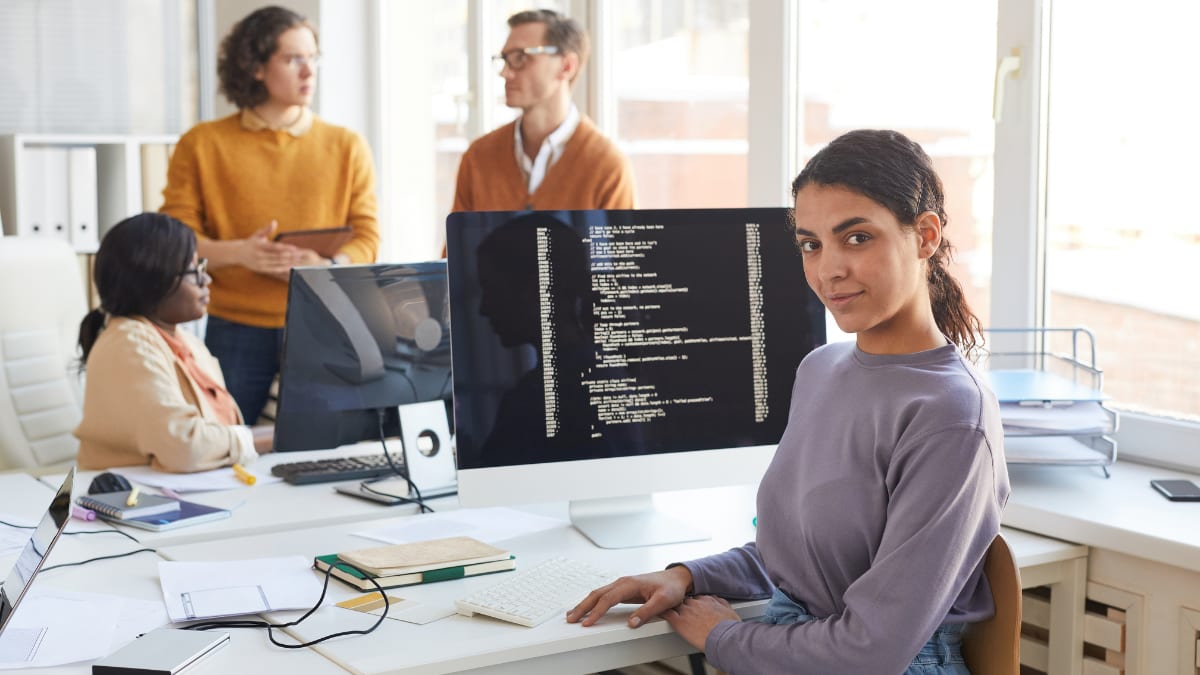 software programmer sitting at a desk in an office with teammates meeting behind her