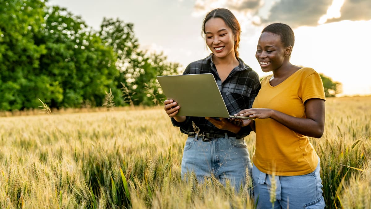 two young agricultural business owners standing in a field holding a laptop