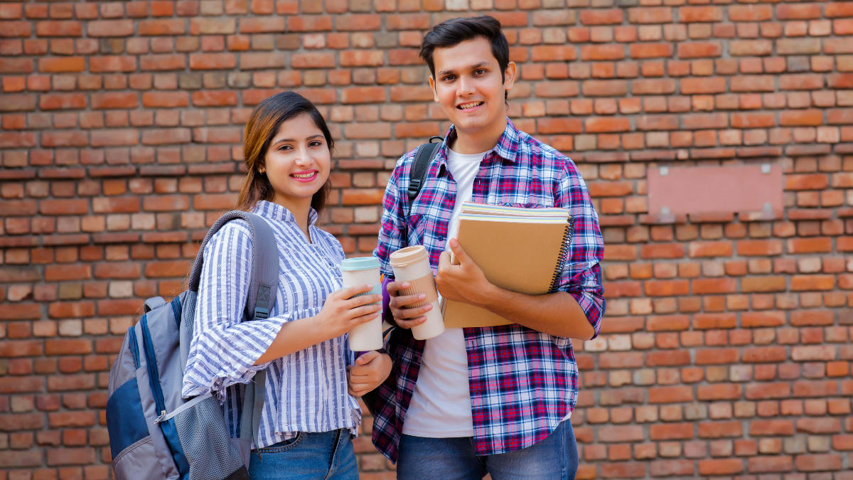 two students holding coffee cups standing against a brick wall