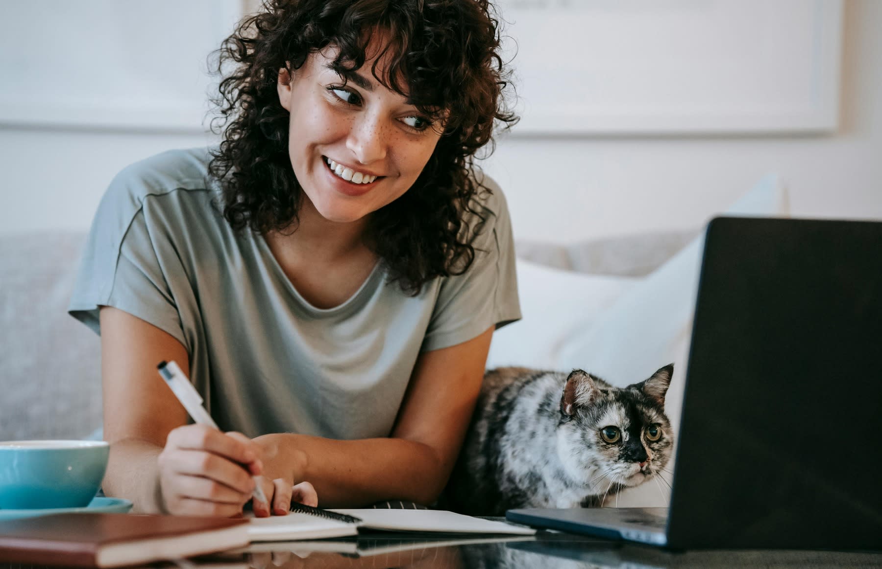Woman writing notes as she looks on her laptop, with her cat beside her
