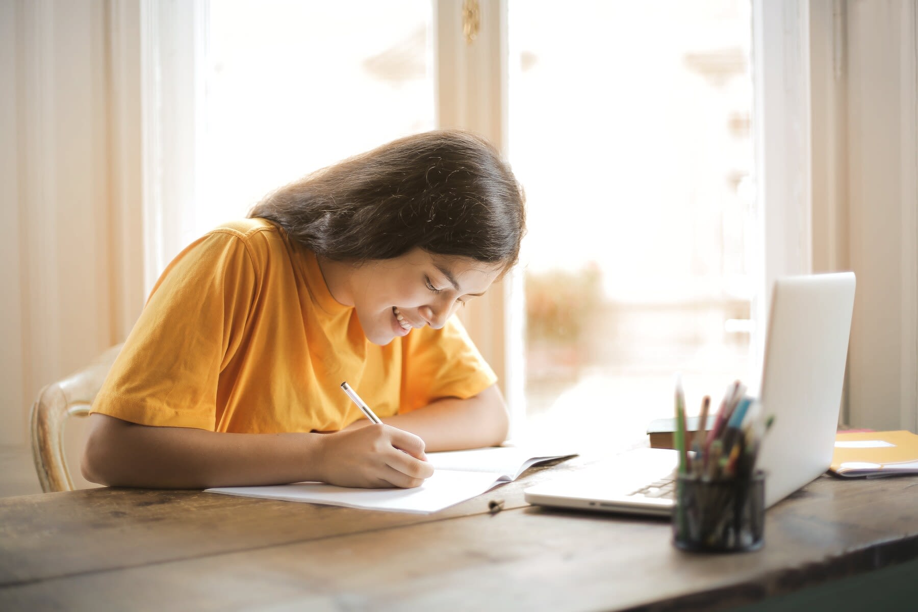 Woman in a yellow shirt writing in a thin notebook on a wooden table with a laptop on it