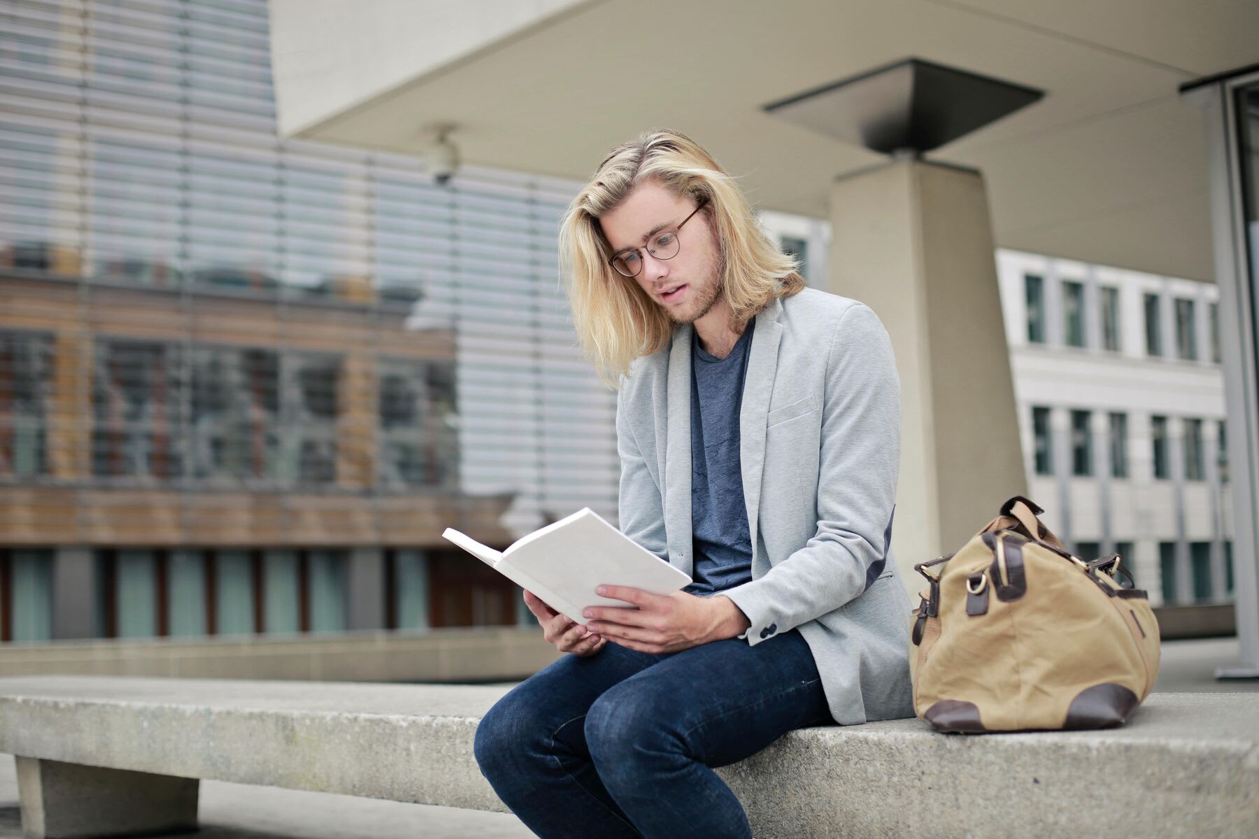Man with long blonde hair and a sports coat sitting on a concrete wall reading a book outside