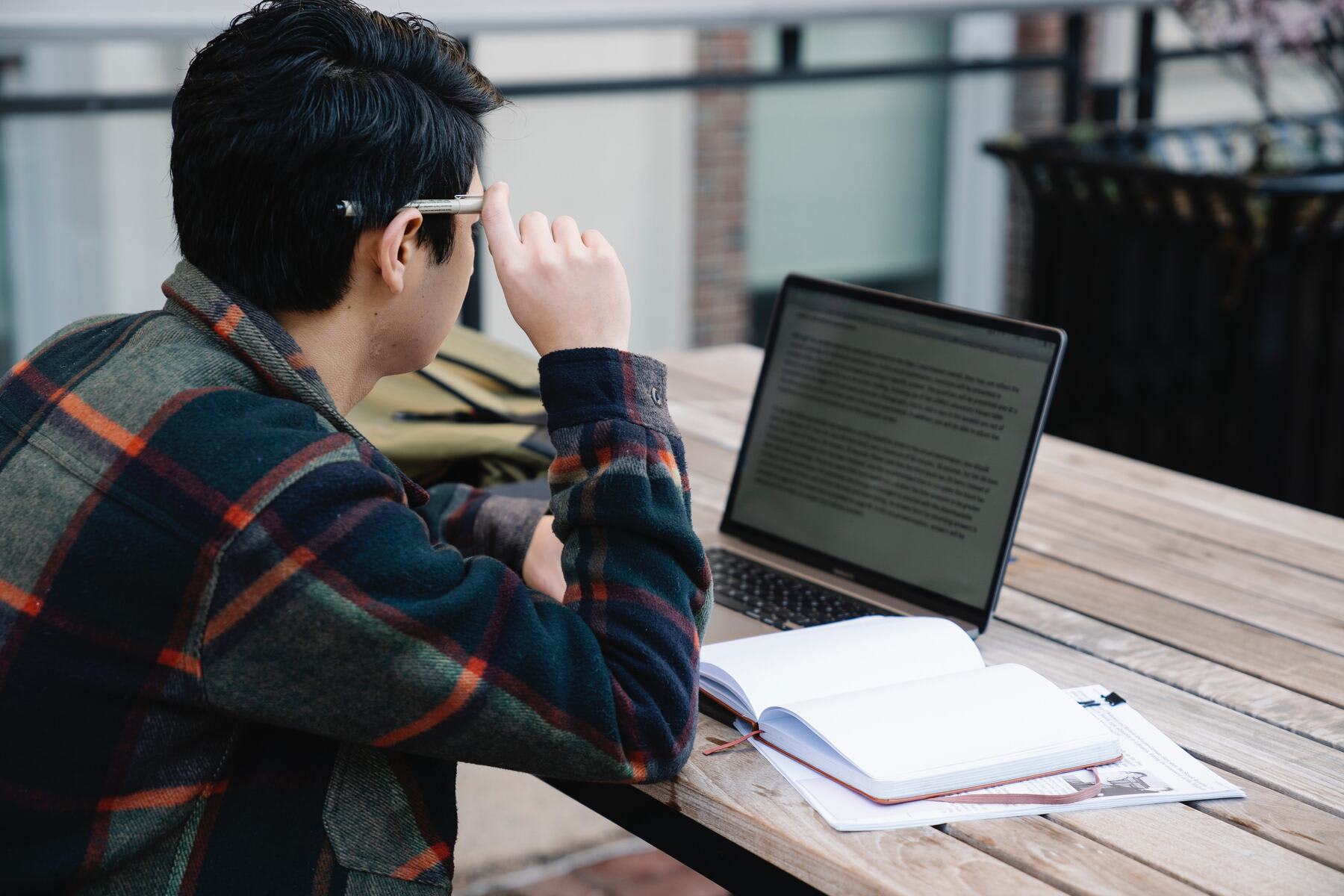 College student holding a pen by his ear and looking at his laptop while sitting at a wooden table outside