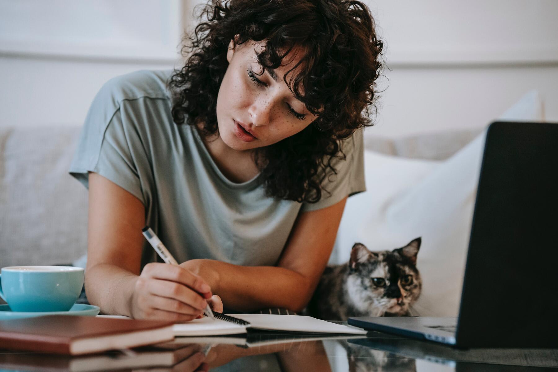Woman writing notes on her notebook while her laptop and cat is beside her