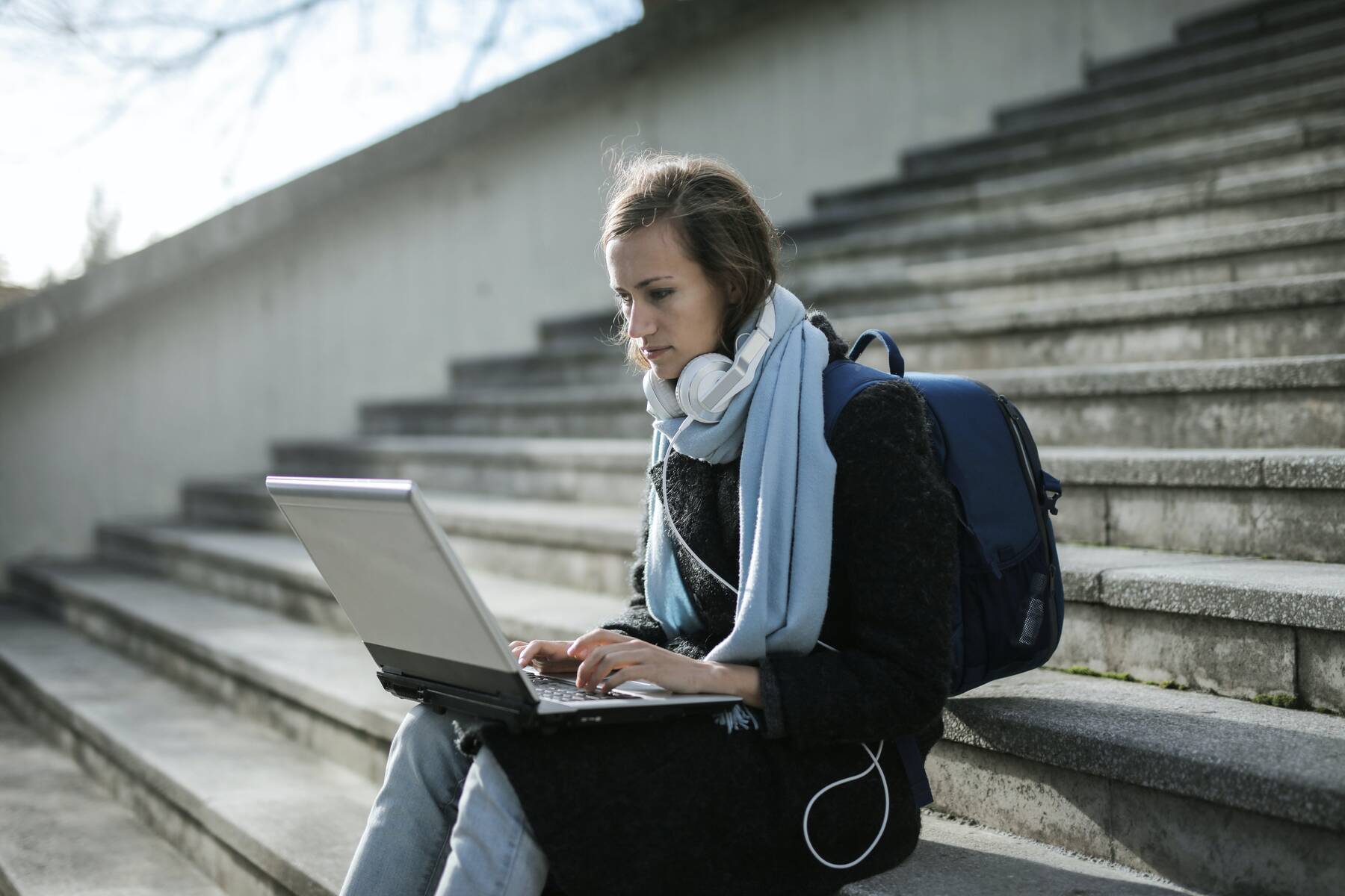 Woman sitting on the steps, while using her laptop and wearing her backpack and headphones