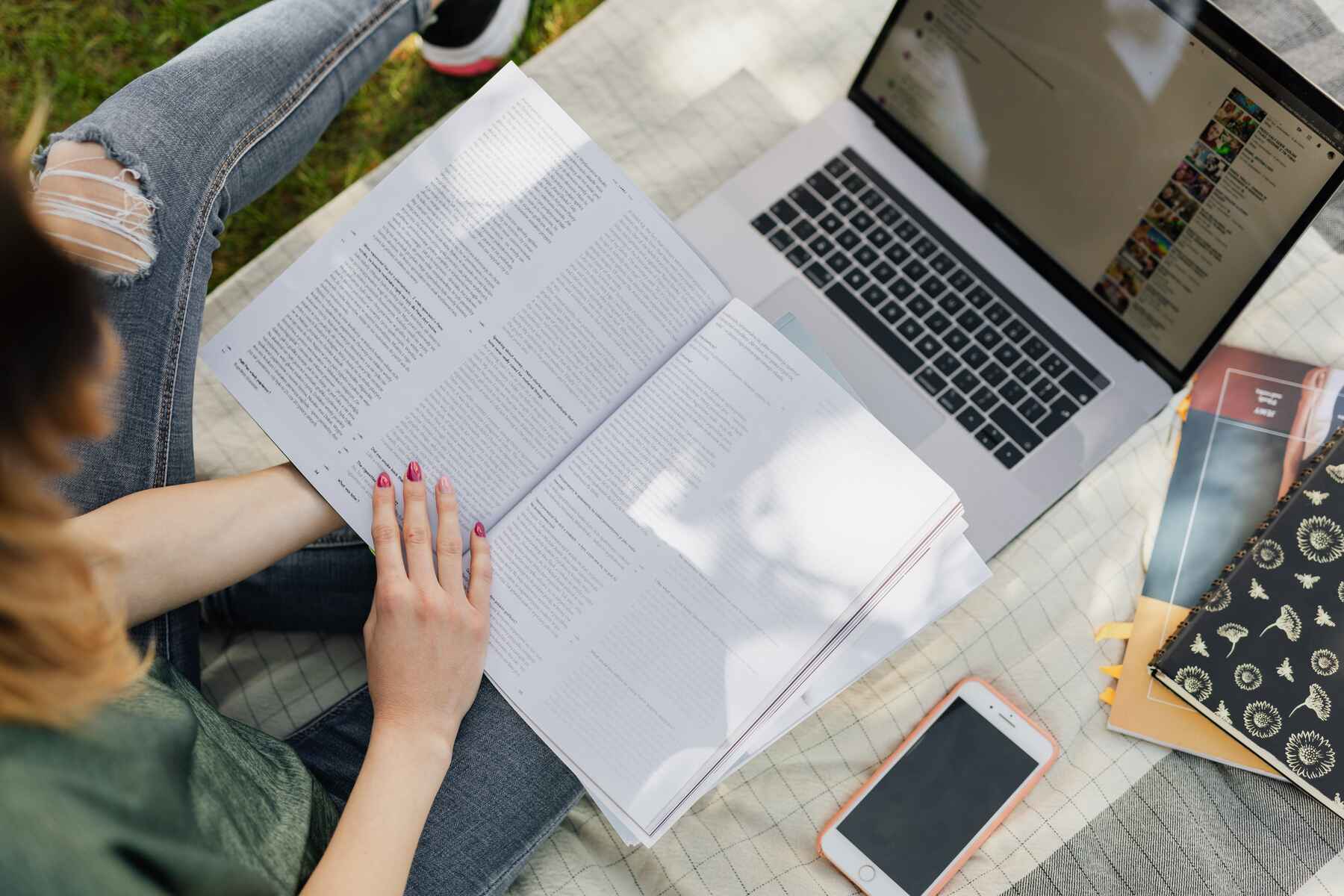 Woman sitting on the grass outdoors and reading a book while her laptop is in front of her