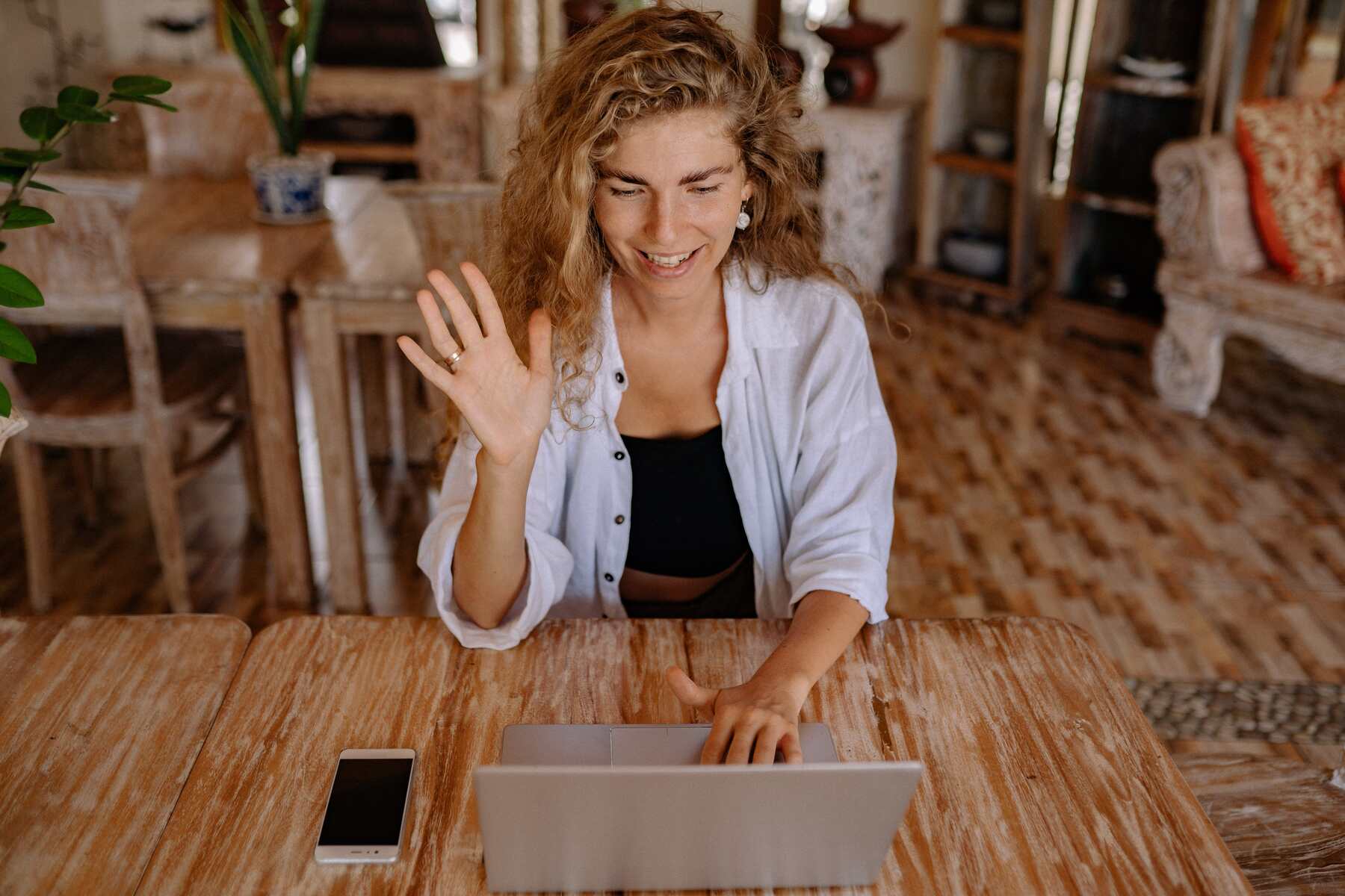 Woman smiling and waving at her laptop screen while sitting at a wooden table in a living room