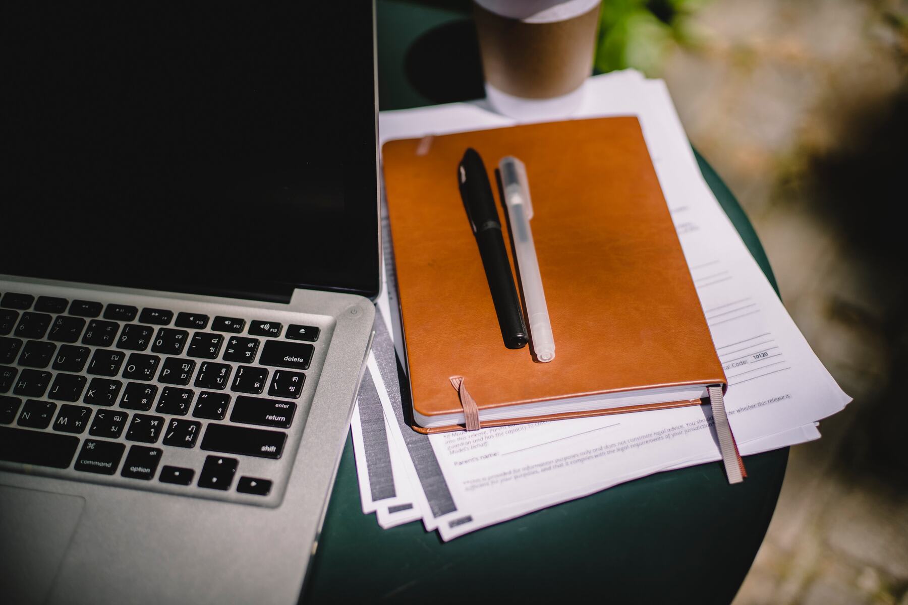 Documents, notebook and pens beside a laptop