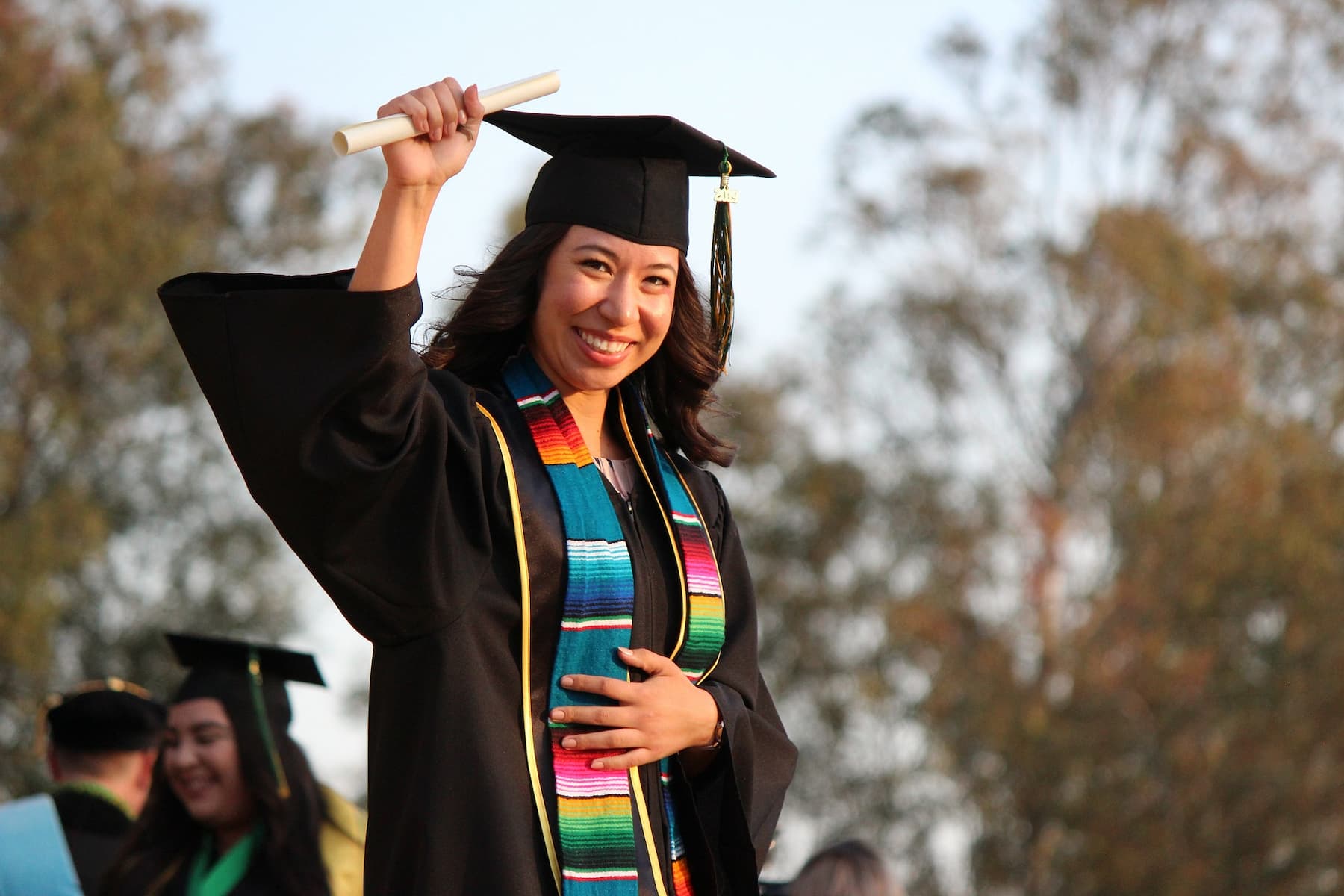 Woman raising her hand to show off her diploma while wearing graduation robes and cap