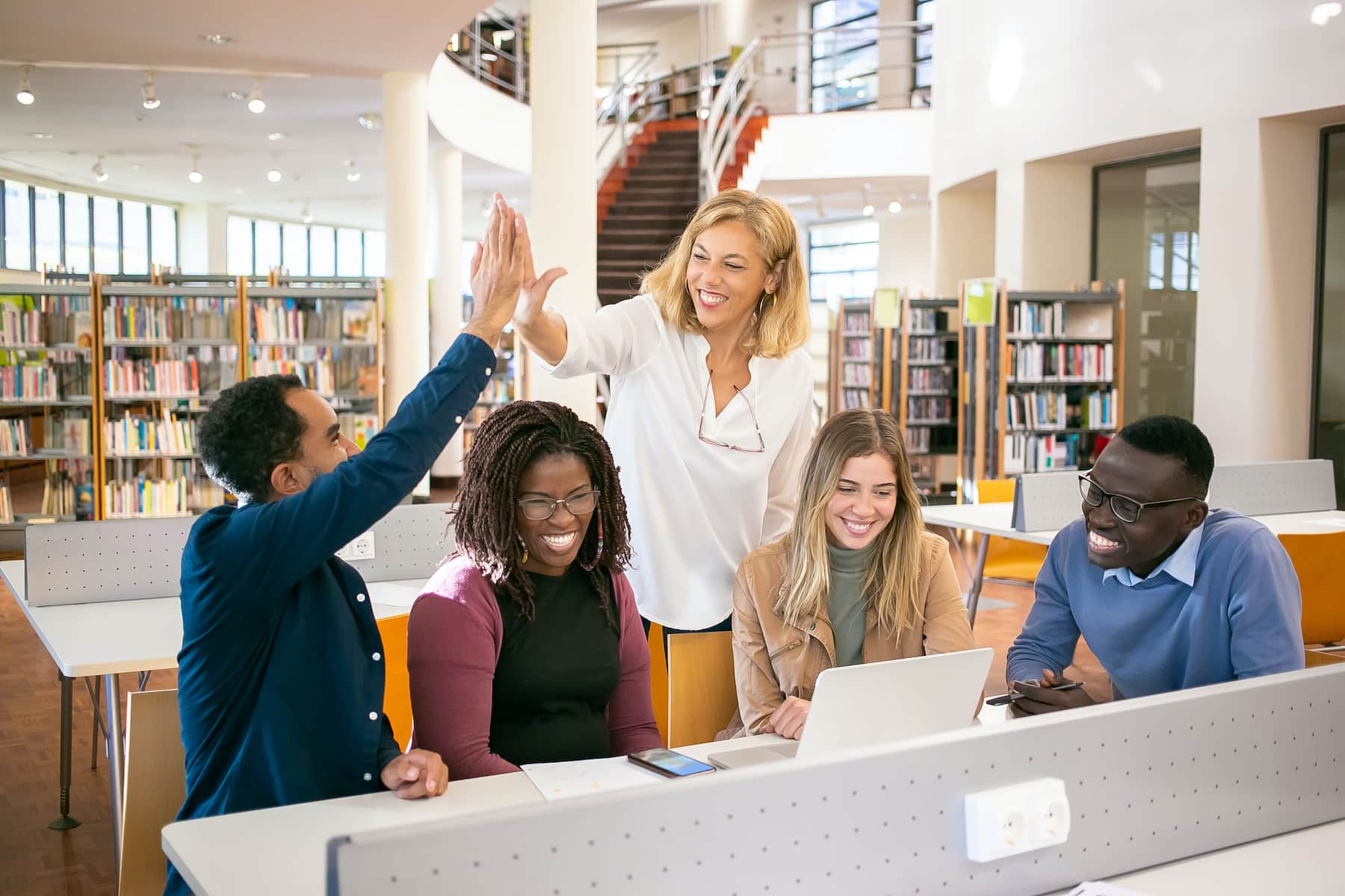 Four students sitting on a long table, while one of them is high fiving a teacher