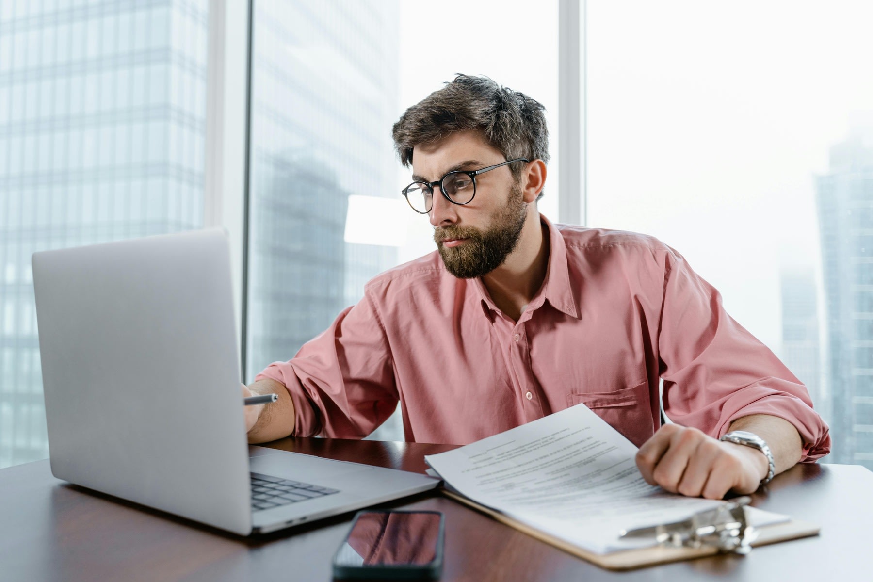 Man looking at his laptop while reviewing printed reports on a clipboard