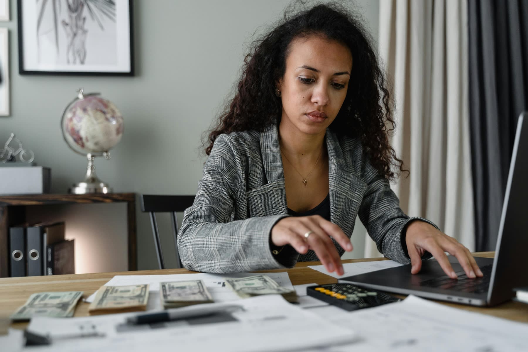 Woman using her calculator and laptop, while dollar bills and printed reports are covering her whole table