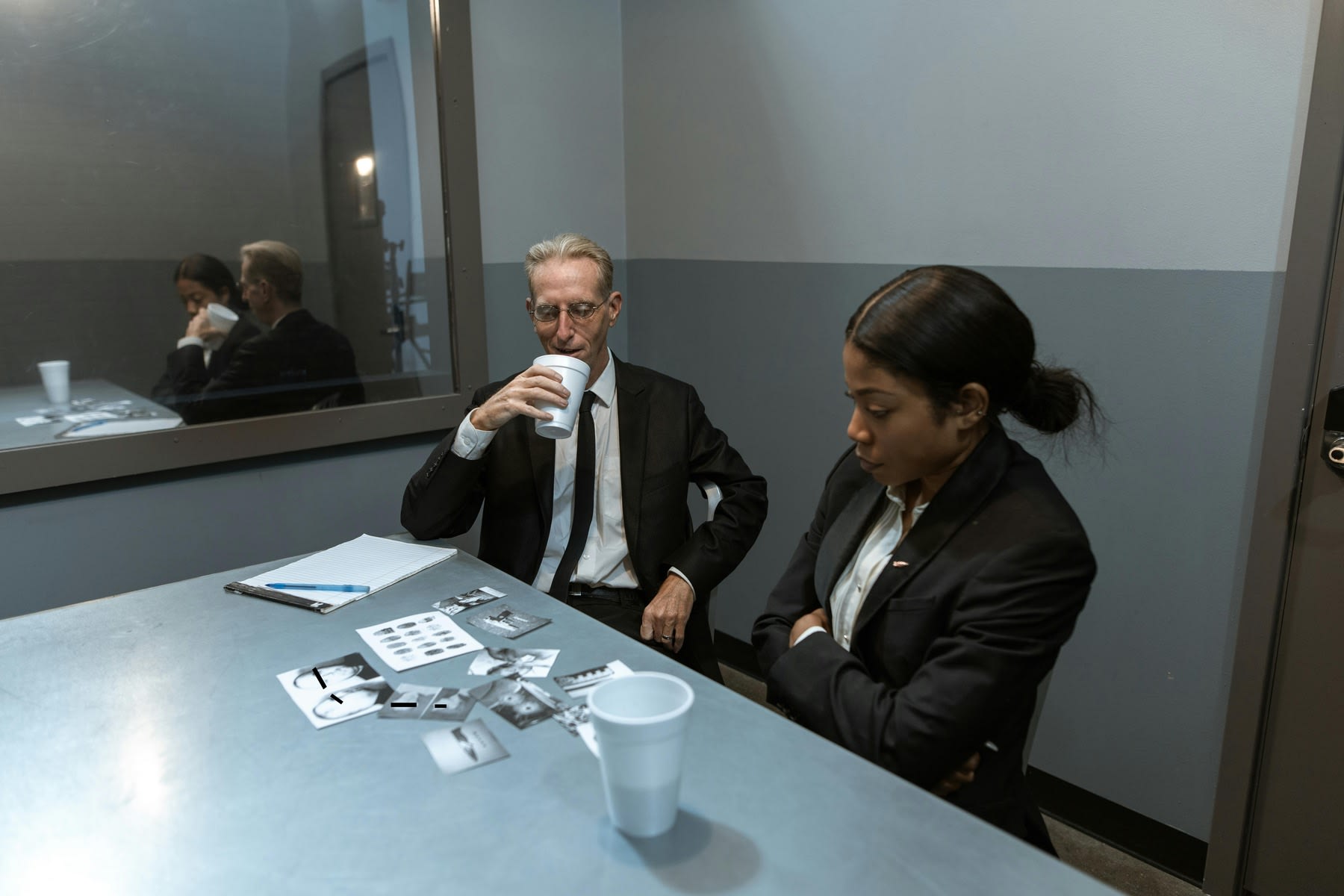 A man and a woman wearing suits, looking at images of suspects in the interrogation room