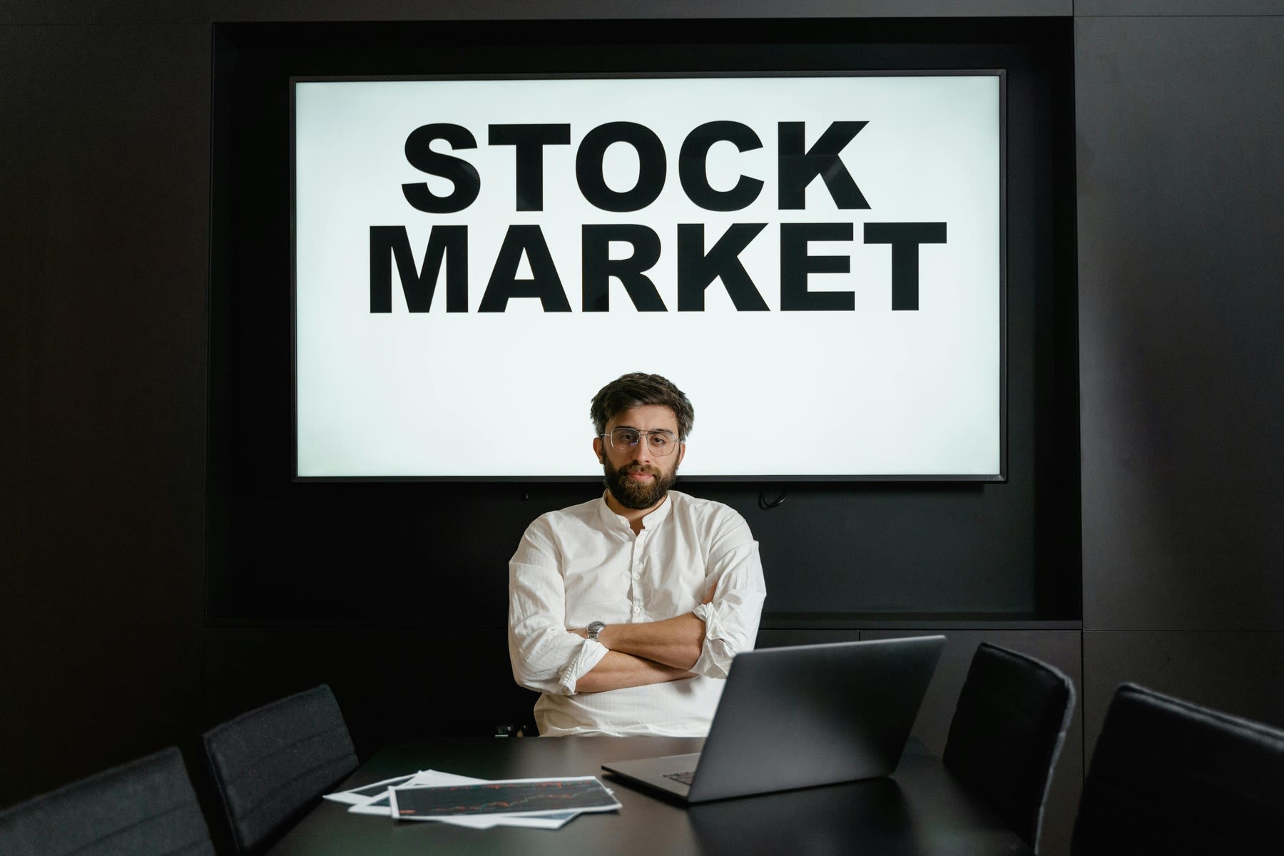 Man looking at his laptop while a huge screen with the text Stock Market is seen behind him