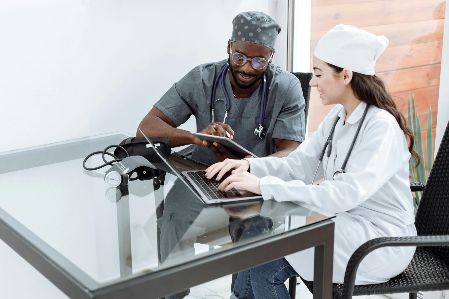 Two people wearing medical scrubs, conversing while typing and taking notes