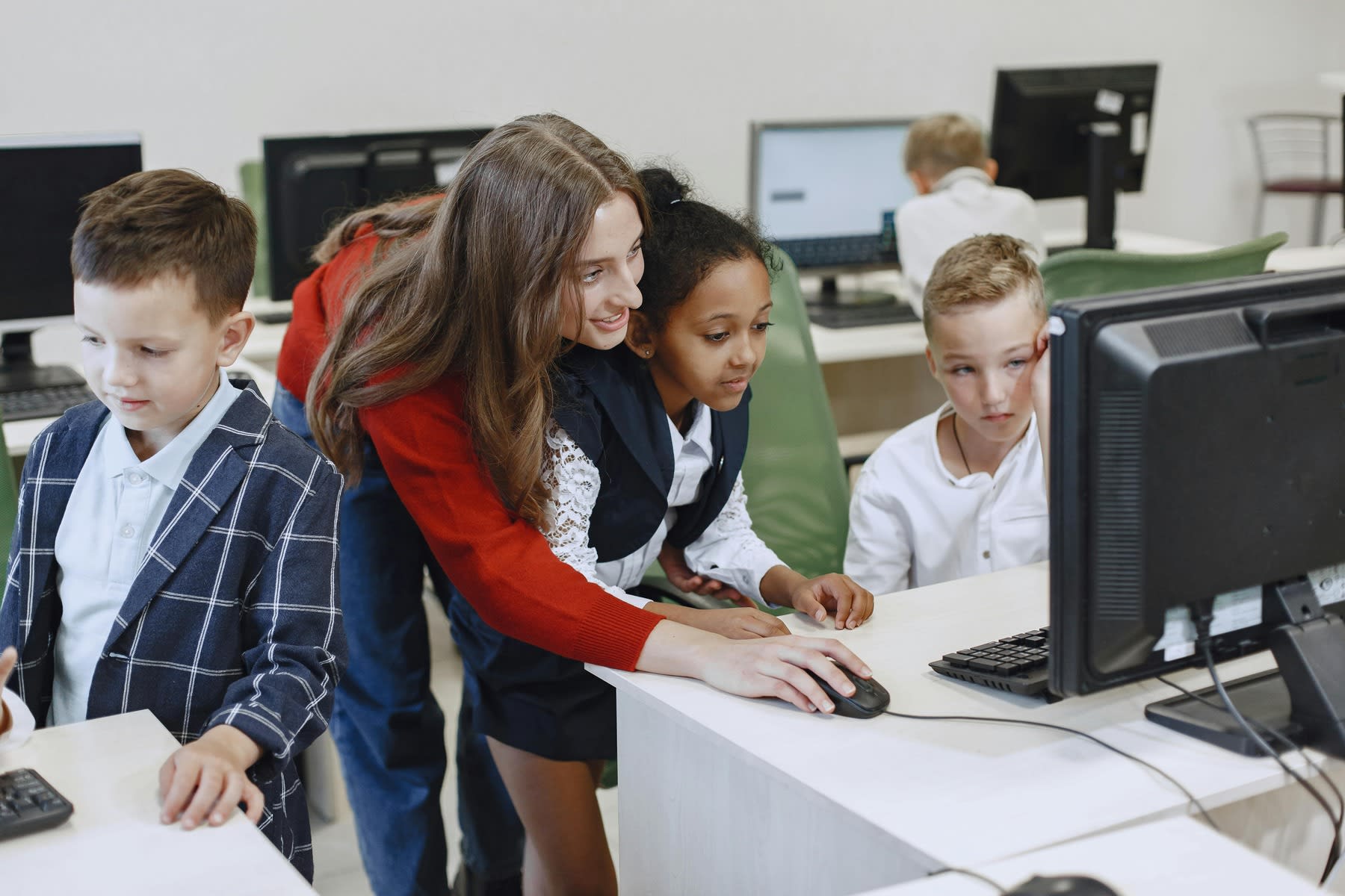 Woman assisting young students as they use desktop computers on a classroom