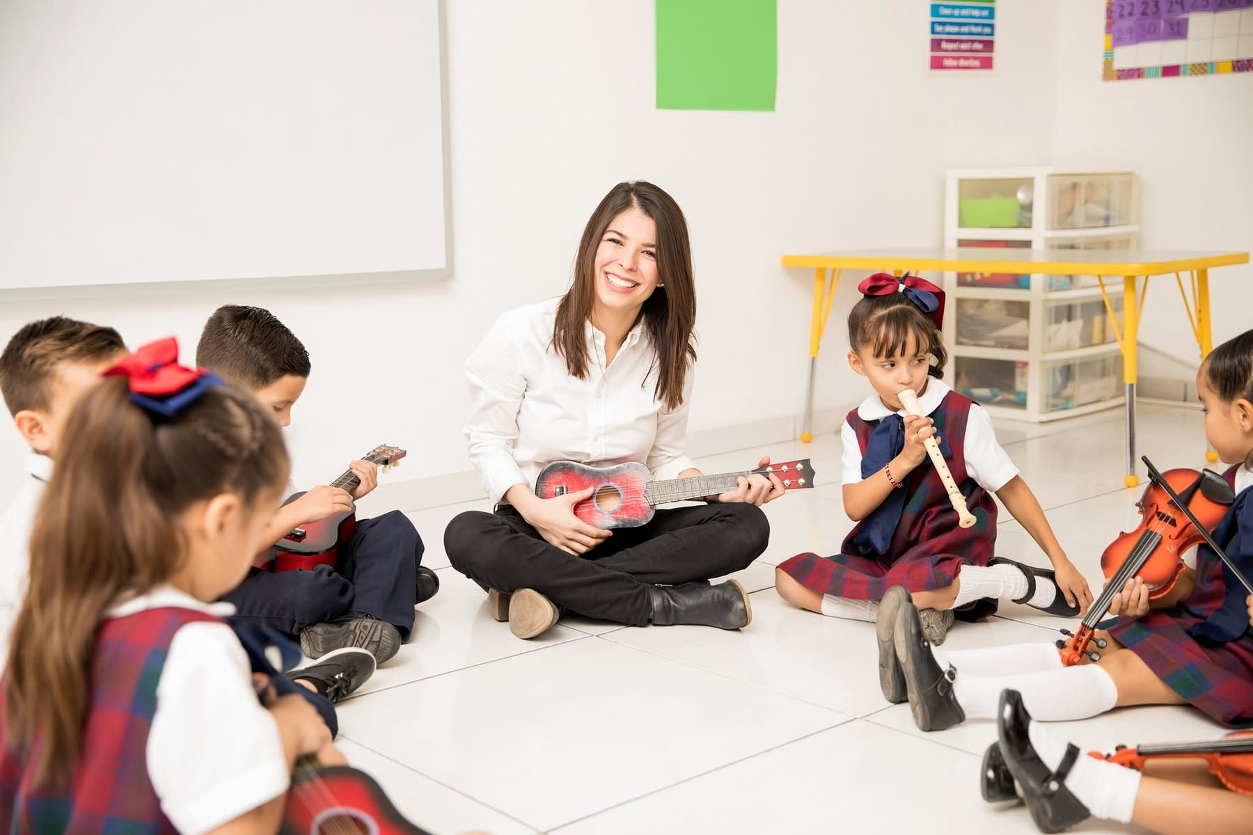 Female teacher playing a small guitar while her kindergarten class are seated in a circle with her