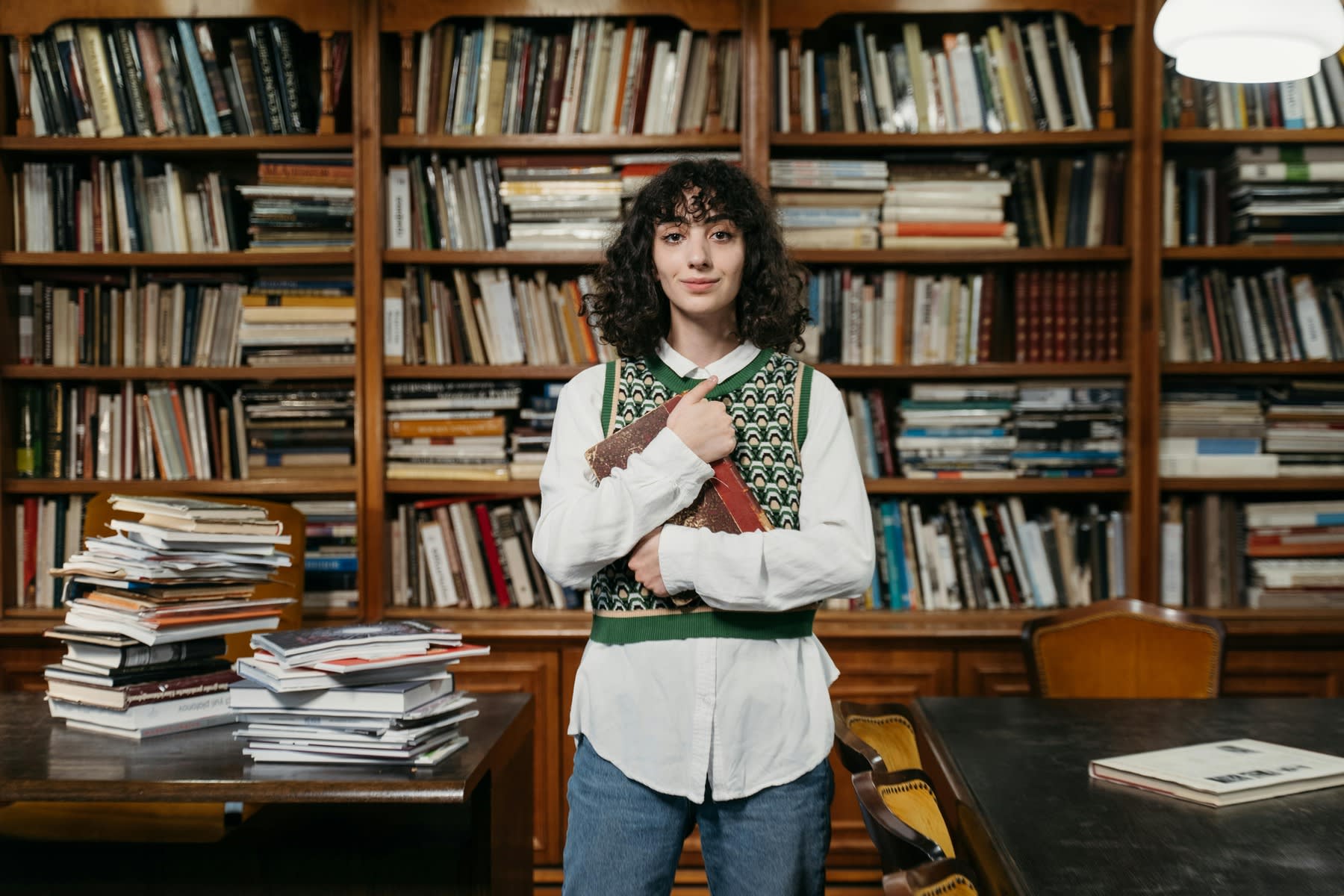 Woman holding a textbook while standing in a room with shelves and tables filled with books