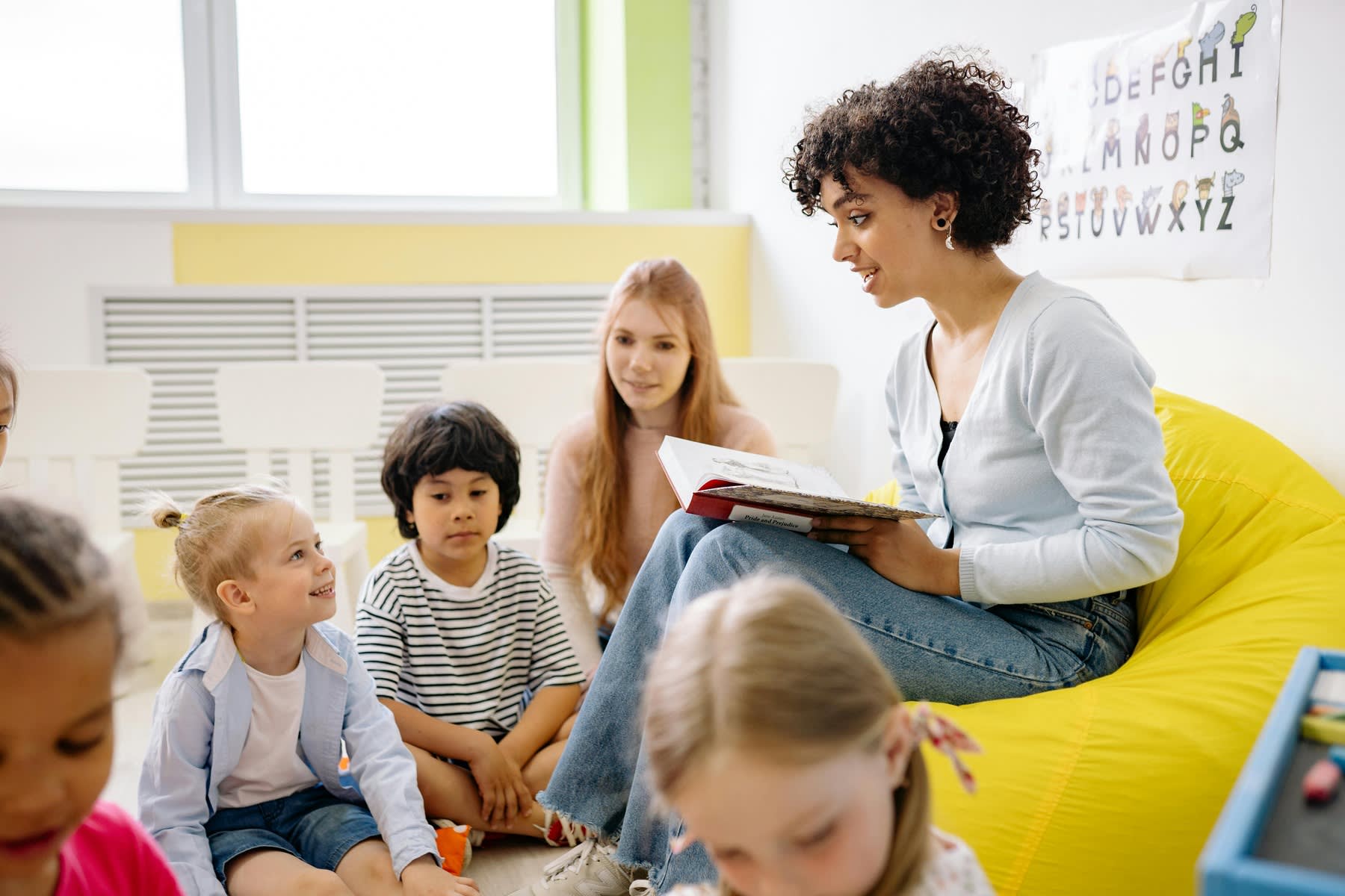 Woman sitting on a bean bag and reading from a book, while her young students are listening