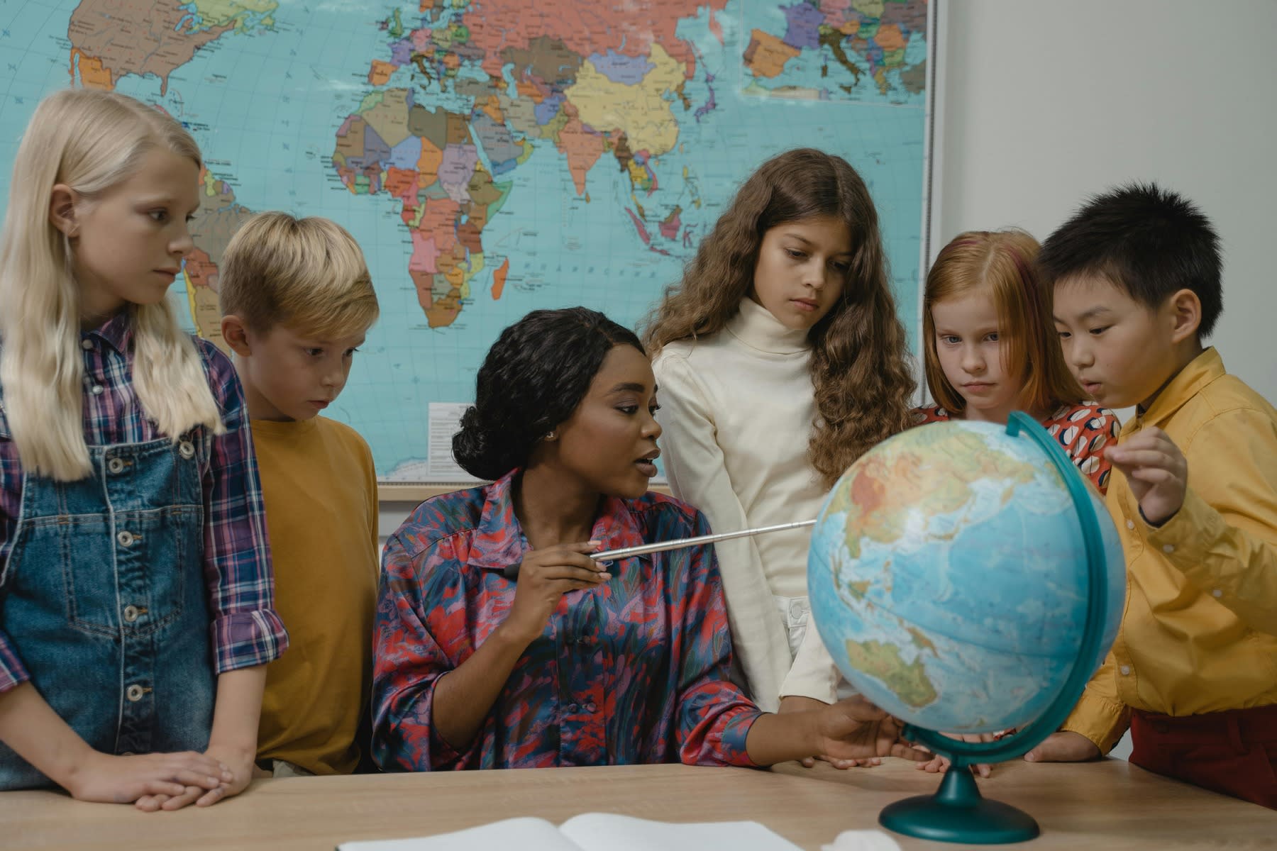 Female teacher pointing at a globe in her desk while her young students looks on