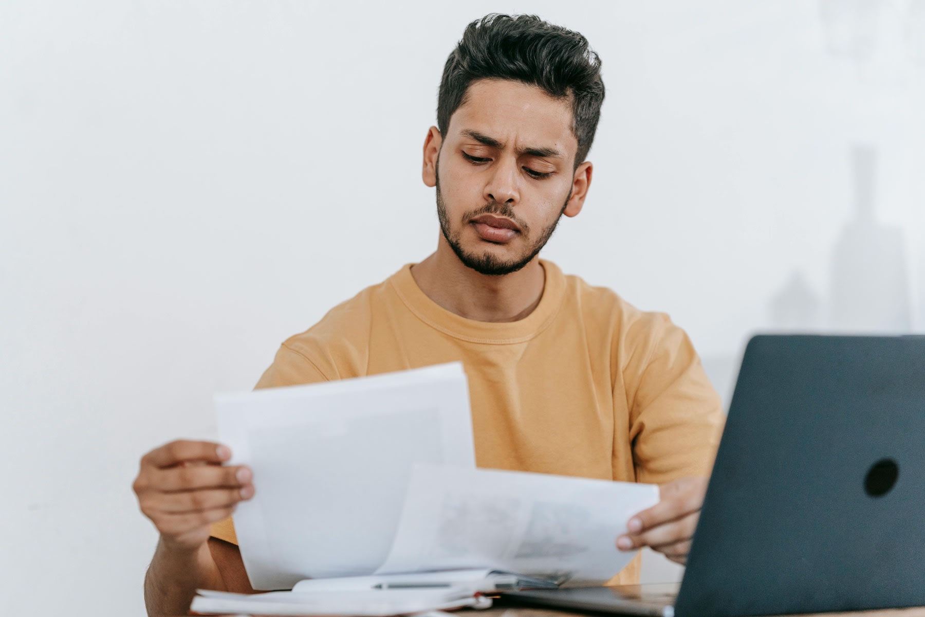 Man reading printed documents with his laptop in front of him