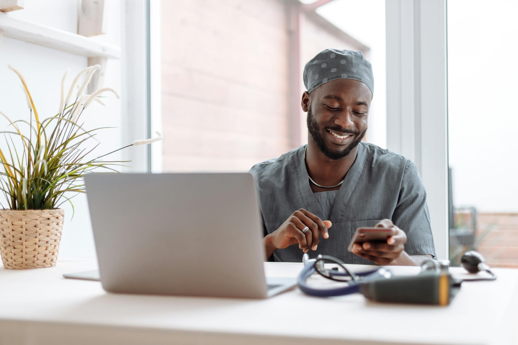 Male nurse looking at his smartphone while sitting in front of a laptop