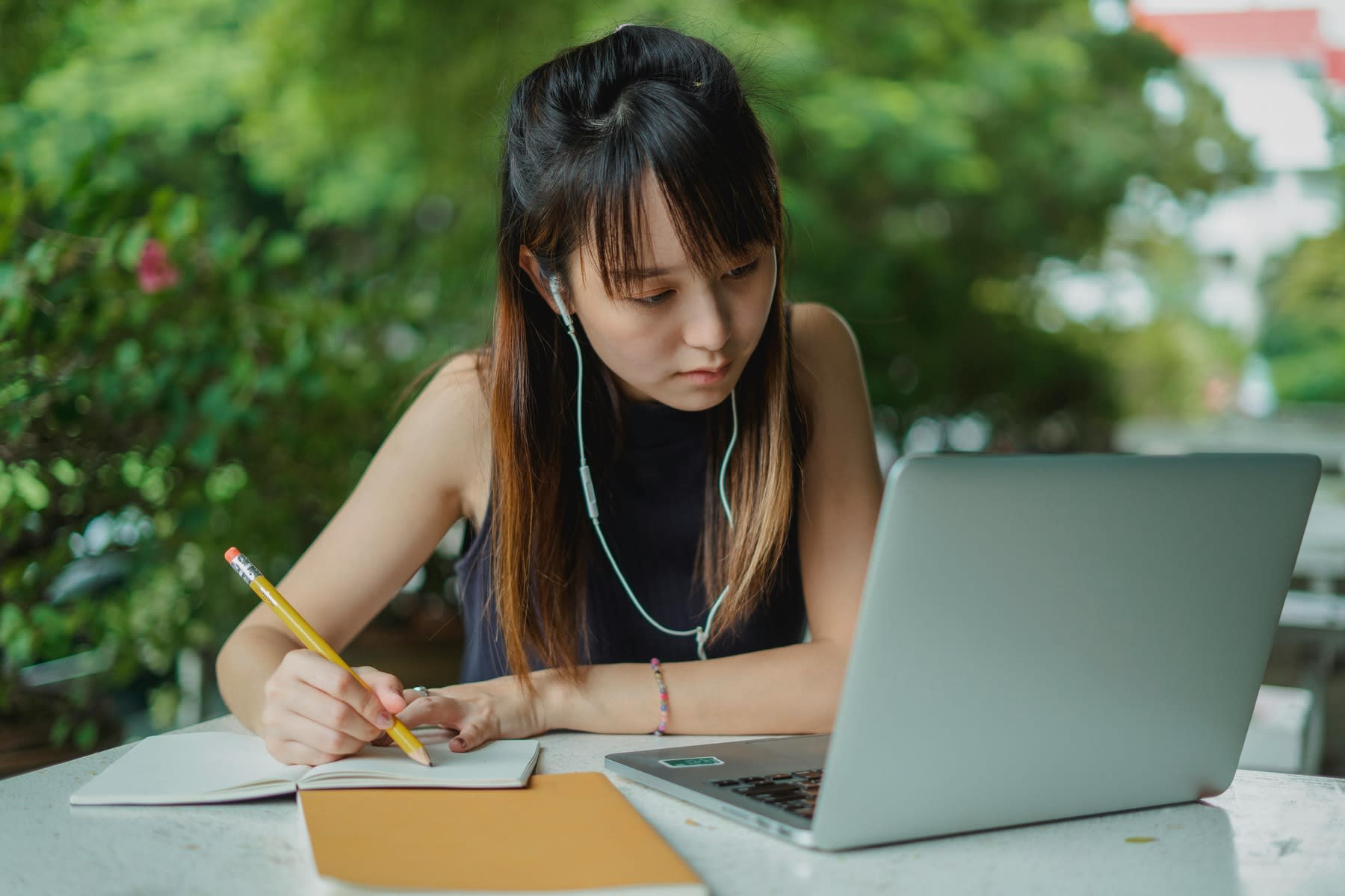 Woman using earphones while looking at her laptop and writing down notes