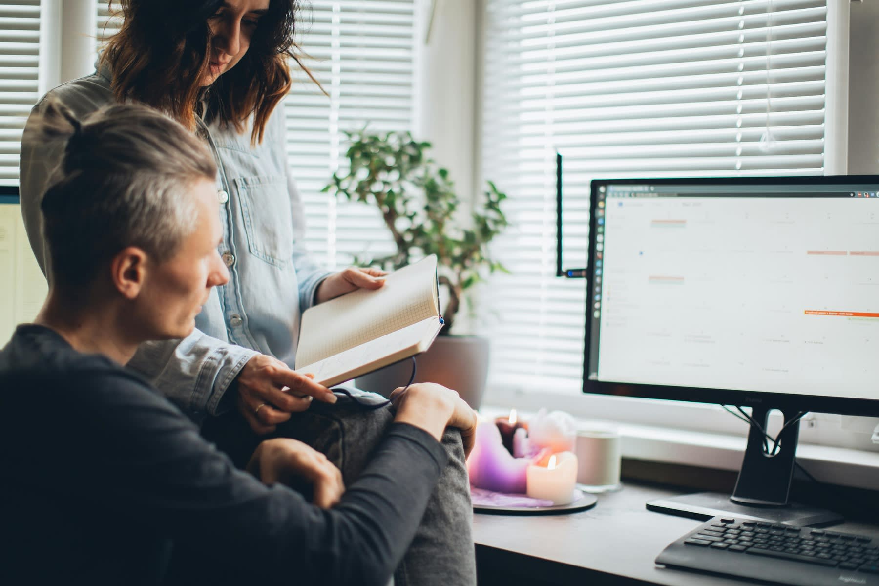 A woman holding a notebook and giving to a man sitting in front of a computer monitor