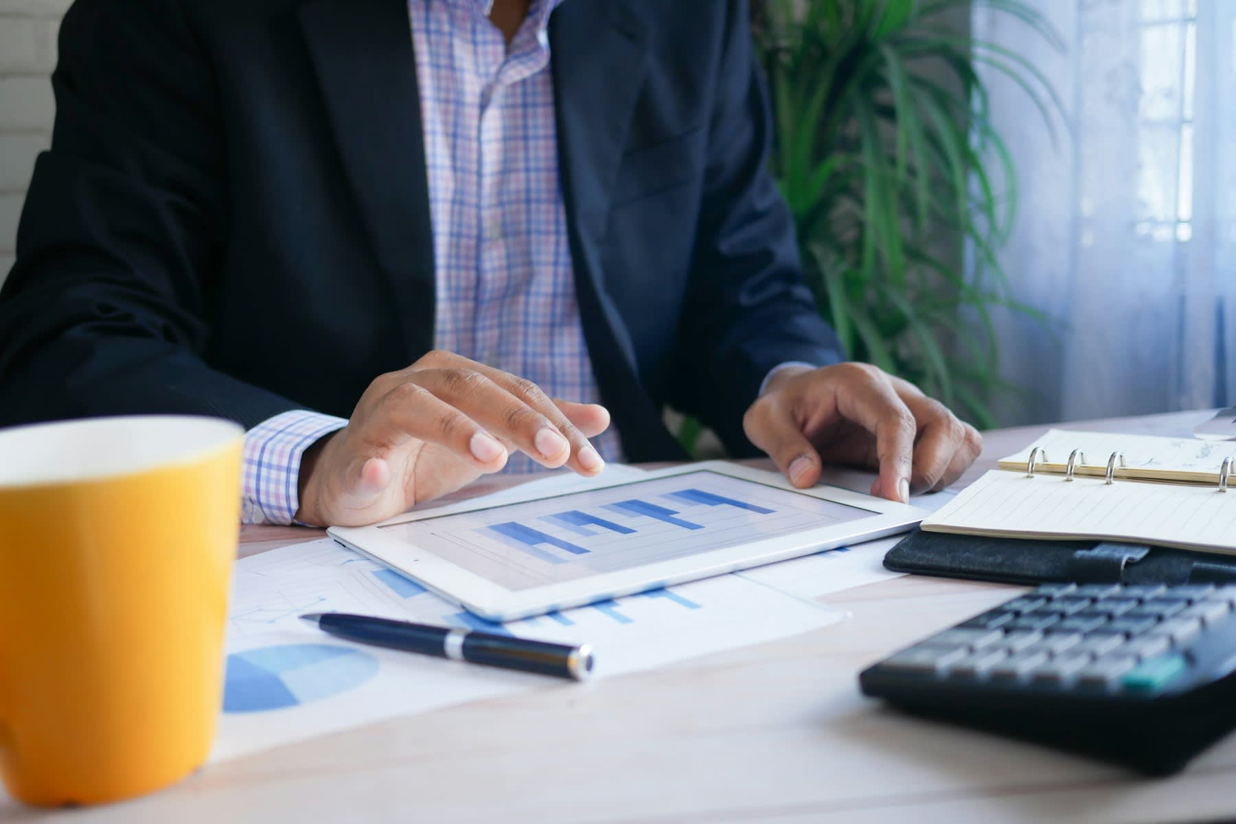 Man looking at data reports on his tablet, while his calculator, printed reports and notebook is on his table