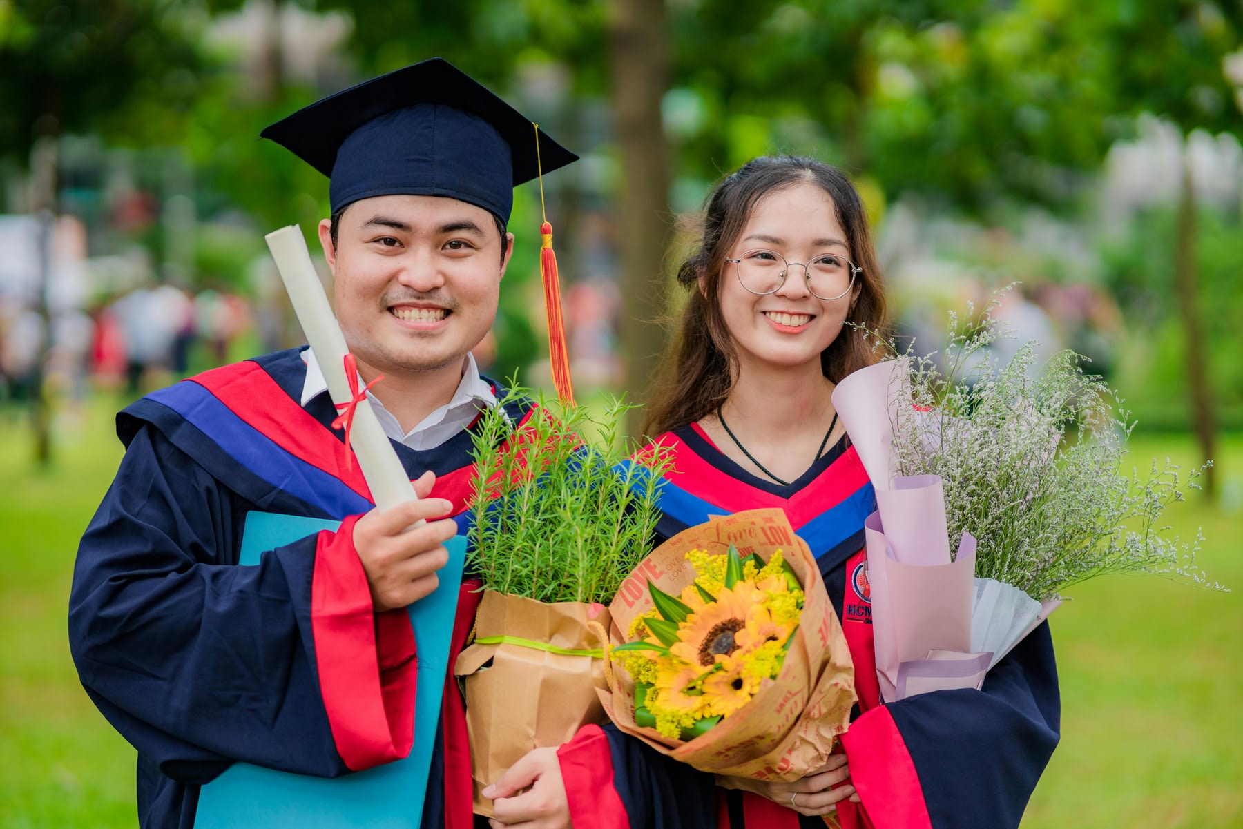 Two students wearing graduation robes, posing for pictures while both holding congratulatory flowers