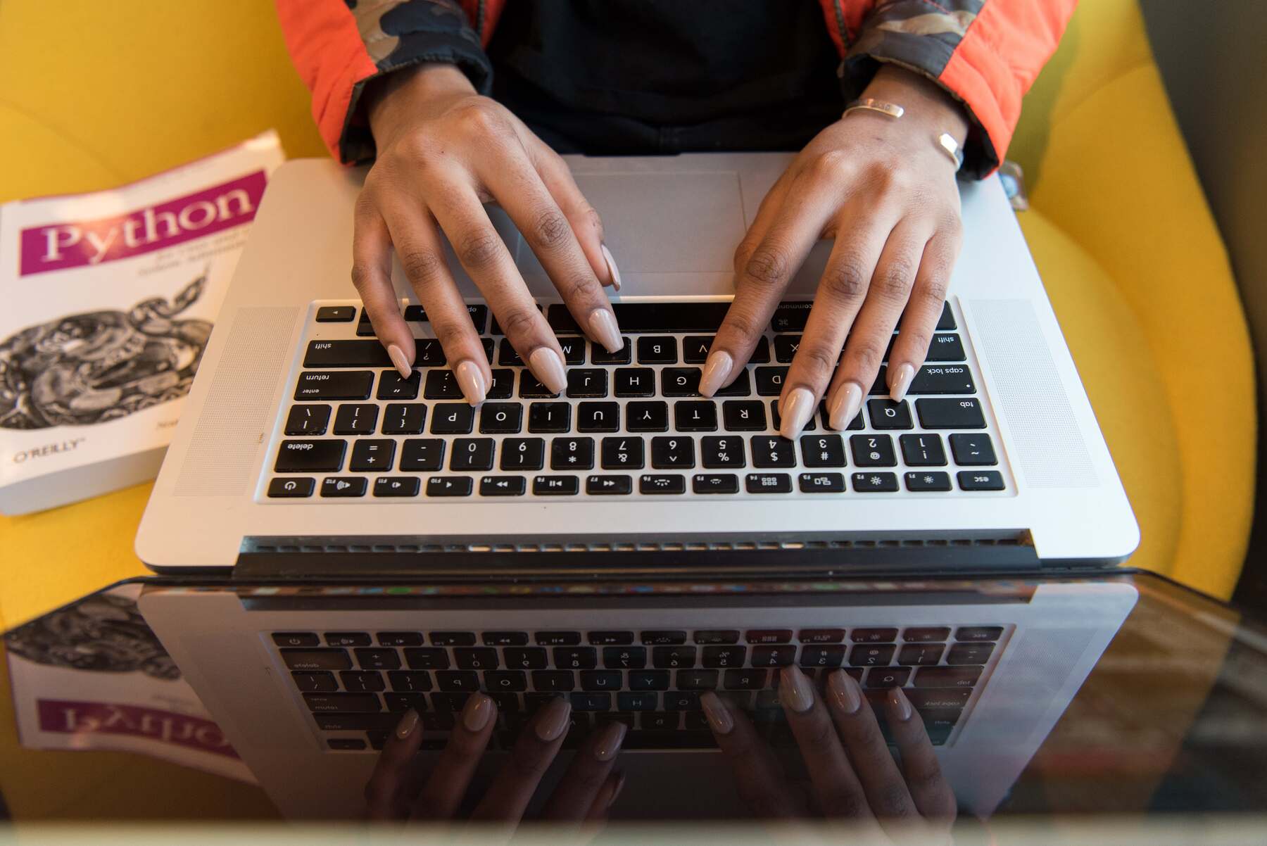 Woman with tan acrylic painted nails typing on a laptop with a python book next to her