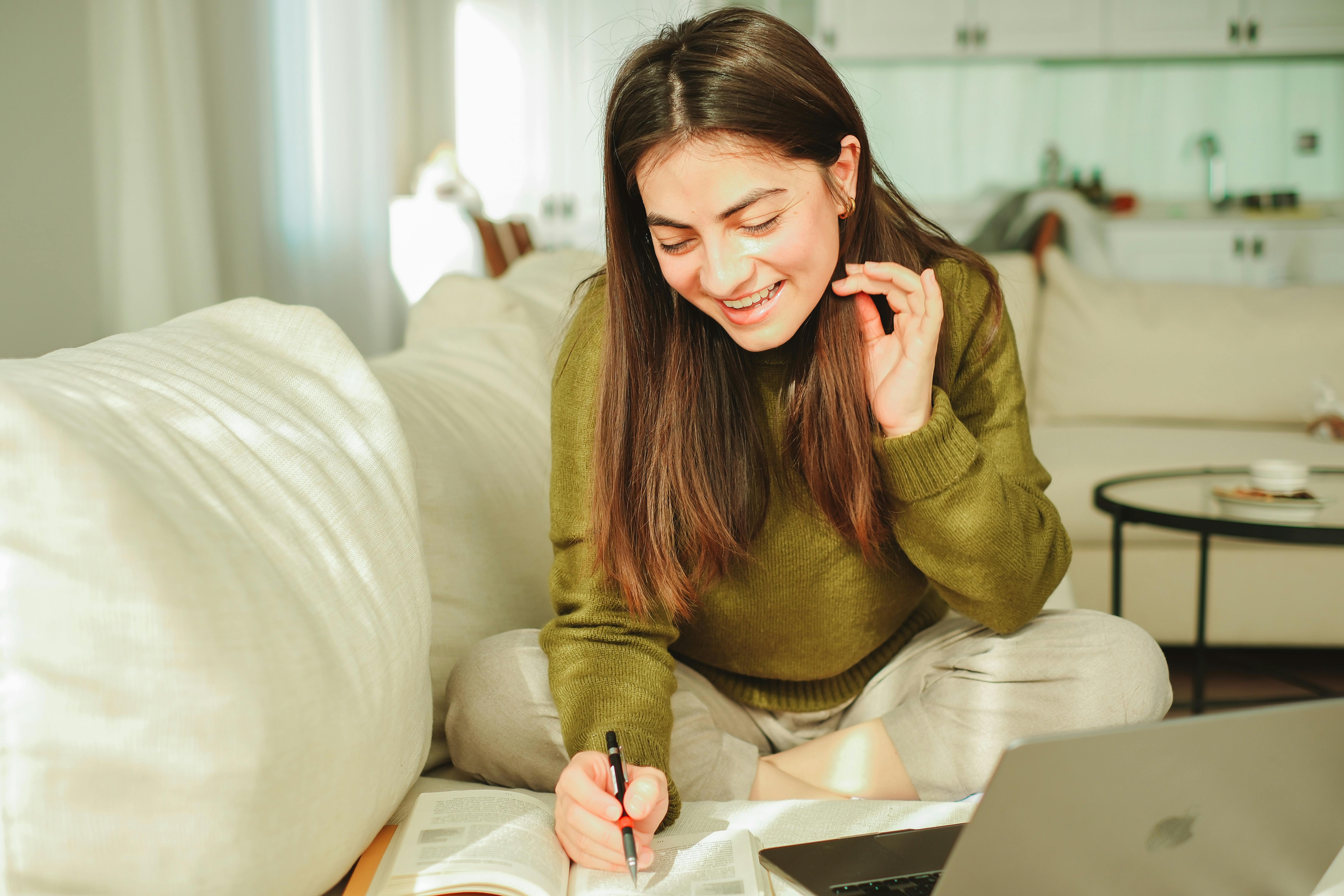 Woman sitting crosslegged on the sofa while writing down notes, with her laptop in front of her
