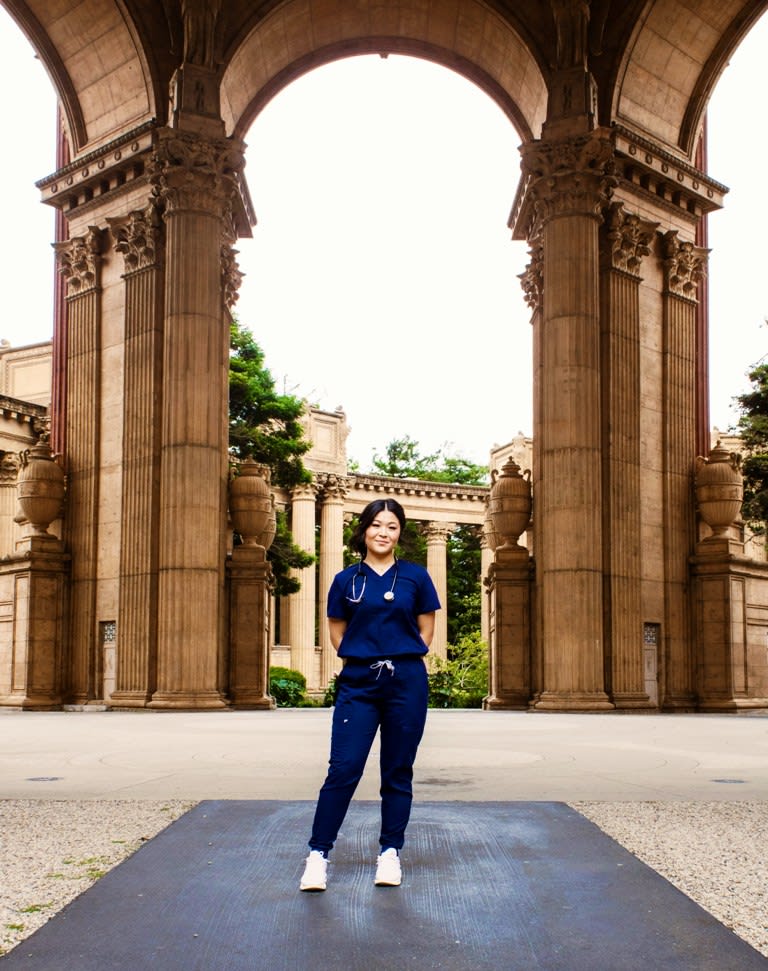 Woman wearing blue nursing scrubs, posing for a photo right behind an outdoor architecture structure